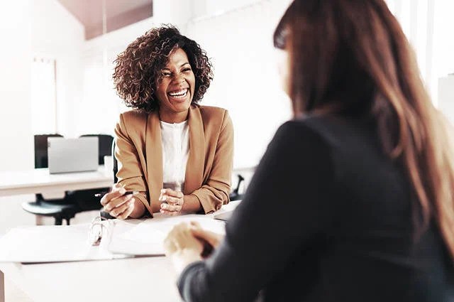 Two women are sitting at a desk in an office, engaged in a friendly conversation. One woman is smiling and holding a pen, while the other is facing away from the camera. Papers and folders are on the desk.