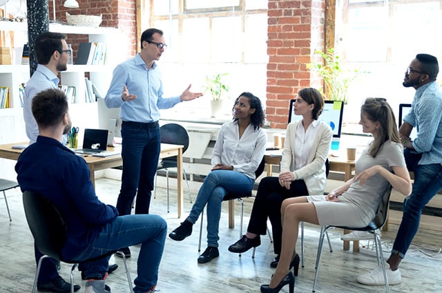 A group of six people sit in chairs while one person stands, speaking and gesturing in a bright office with brick walls and large windows. The group appears engaged in a team discussion or meeting.