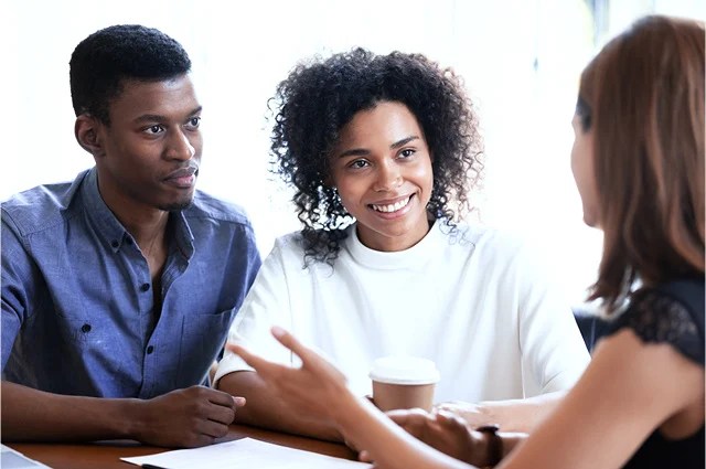 A man and a woman sit together at a table, smiling and listening to another woman who is speaking to them. The woman in the center holds a coffee cup. They appear engaged in a friendly conversation or meeting. A man and a woman sit together at a table, smiling and listening to another woman who is speaking to them. The woman in the center holds a coffee cup. They appear engaged in a friendly conversation or meeting.