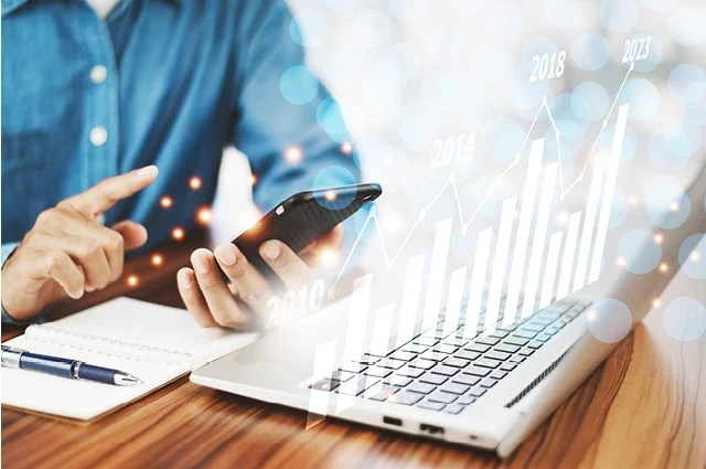 A person using a smartphone and laptop, with a notebook nearby. Overlaid are upward-trending bar and line graphs labeled with years from 2019 to 2023, symbolizing business growth and technology. A person using a smartphone and laptop, with a notebook nearby. Overlaid are upward-trending bar and line graphs labeled with years from 2019 to 2023, symbolizing business growth and technology.