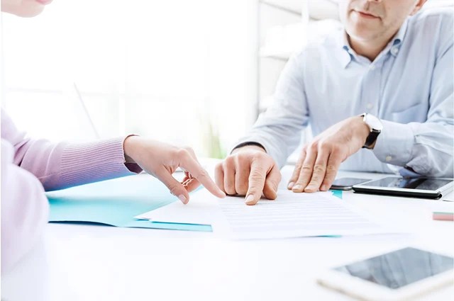 Two people sit at a desk, reviewing and pointing at a document together. The scene includes papers, a tablet, and a smartphone, suggesting a professional or office environment. Faces are partially out of frame.