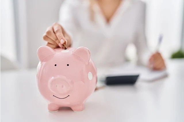 A person in a white shirt places a coin into a pink piggy bank on a table, with a calculator and notepad in the background. A person in a white shirt places a coin into a pink piggy bank on a table, with a calculator and notepad in the background.