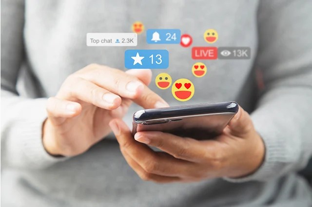 A person uses a smartphone, with social media notifications and reaction icons such as likes, hearts, and emojis digitally overlaid above the screen.