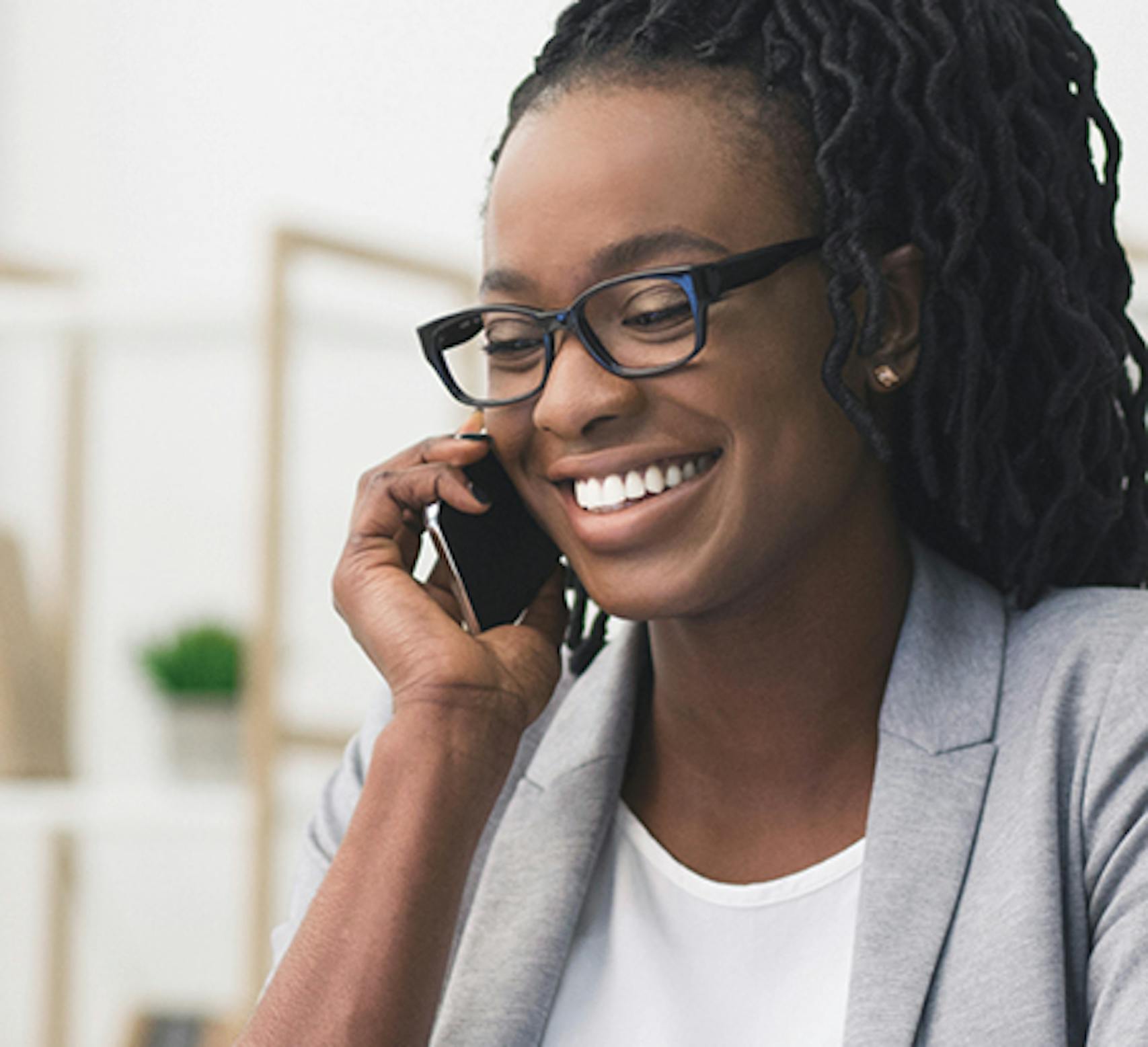 A woman with glasses and long braided hair smiles while talking on a smartphone. She is wearing a light gray blazer and a white top, sitting in a bright office environment. A woman with glasses and long braided hair smiles while talking on a smartphone. She is wearing a light gray blazer and a white top, sitting in a bright office environment.