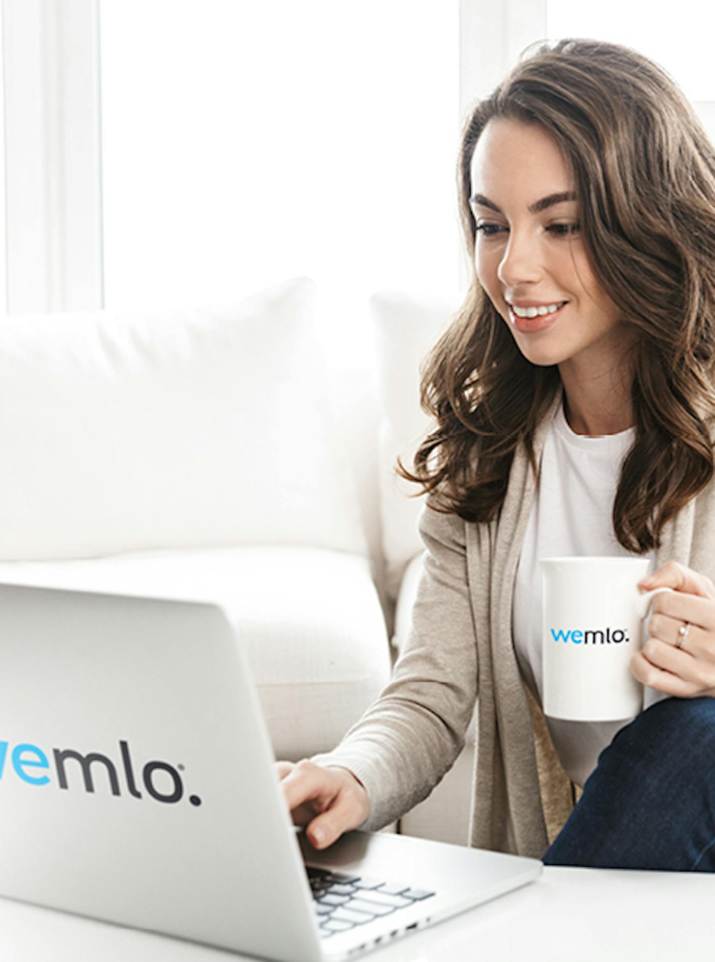 A woman sits on a white sofa, smiling while working on a laptop with wemlo. logo. She holds a white mug, also labeled wemlo., in a bright, modern living space.