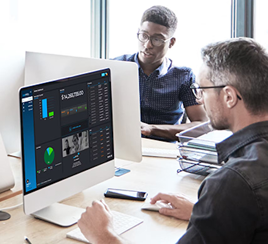 Two men work at a desk with desktop computers displaying data analytics dashboards and charts. One man focuses on his screen while the other looks toward him, suggesting collaboration in a modern office setting.