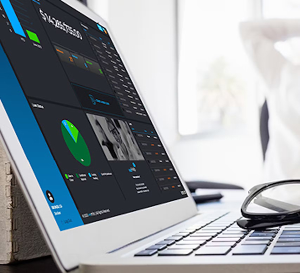 A close-up of a laptop displaying colorful data charts and dashboards. In the background, an out-of-focus person stretches with arms behind their head, and eyeglasses rest next to the laptop.