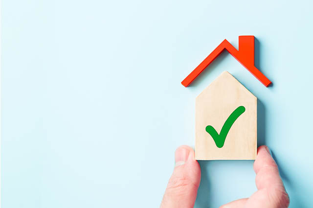 A hand holds a small wooden house with a green check mark, next to a red roof shape on a light blue background, symbolizing approved or safe housing.