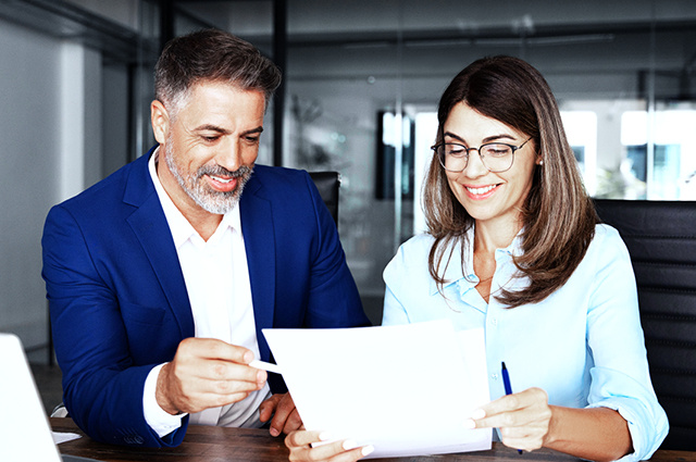 A man in a blue suit and a woman in glasses and a light blue blouse are sitting at a desk, smiling and looking over documents together in a modern office setting.