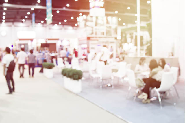 A blurred image of people sitting and standing in a brightly lit indoor event or exhibition space, with tables, chairs, and decorative plants visible.