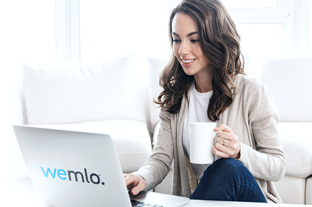 A woman sits on a couch, smiling as she looks at a laptop with “wemlo.” on the lid. She holds a white mug in one hand and appears relaxed in a bright, modern living room. A woman sits on a couch, smiling as she looks at a laptop with “wemlo.” on the lid. She holds a white mug in one hand and appears relaxed in a bright, modern living room.