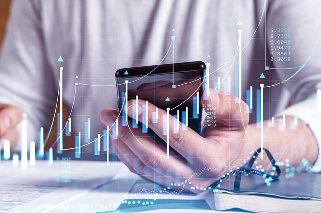 A person holding a smartphone with financial graphs and data overlaid, suggesting analysis or monitoring of stock market trends. A notebook, glasses, and papers are visible on the desk.