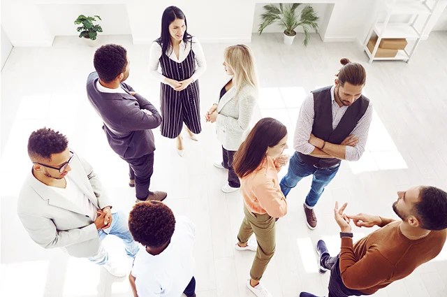 A group of eight adults stand in small clusters, conversing in a bright, modern room with light flooring and plants in the background. They appear to be engaged in friendly discussion.