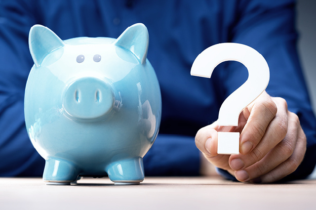 A person in a blue shirt holds a white question mark next to a light blue piggy bank on a table, suggesting uncertainty about savings or financial decisions.