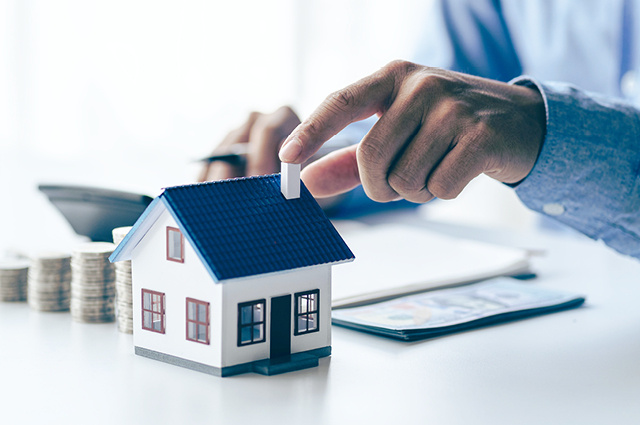 A person’s hand points to the chimney of a small model house on a desk, with stacks of coins, a calculator, and documents in the background, suggesting financial planning or real estate investment.