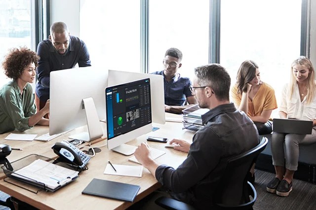 Six people are working together in a modern office. Some are seated at desks with computers, while others are standing or sitting nearby, engaged in discussion and collaboration. Sunlight streams through large windows.
