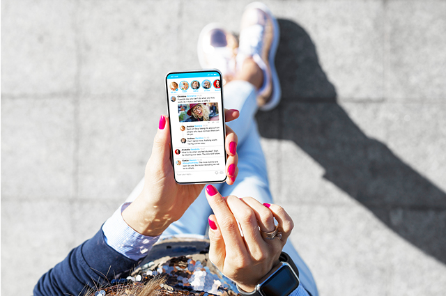 A person with pink nail polish is holding and using a smartphone outdoors, viewing a social media feed. The person is dressed casually in jeans and sneakers, and sunlight casts a shadow on the pavement.