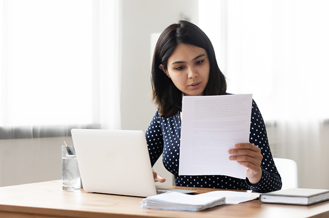 A woman sits at a desk with a laptop, holding and reading a sheet of paper. A glass of water, pen holder, and notebook are also on the table. She appears focused and is wearing a dark polka dot top.