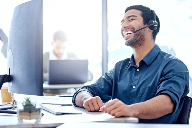 A man wearing a headset sits at a desk, smiling and laughing while looking at his computer screen. Another person is working on a laptop in the background in a bright office setting.