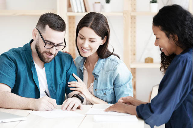 A smiling couple sits at a table as the man signs a document. A woman across from them, also smiling, holds papers and appears to guide them through the process in a bright, modern office setting. A smiling couple sits at a table as the man signs a document. A woman across from them, also smiling, holds papers and appears to guide them through the process in a bright, modern office setting.