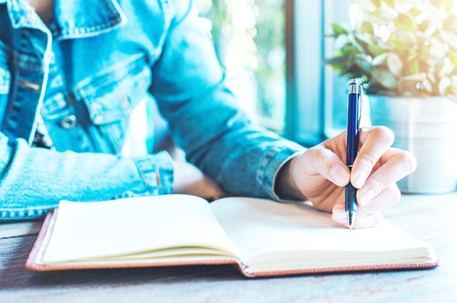 A person wearing a denim jacket writes in an open notebook with a pen at a wooden table near a sunlit window, with a potted plant in the background.