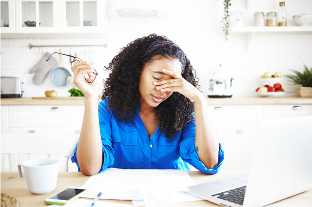 A woman in a blue shirt sits at a kitchen table with a laptop and papers, holding her glasses in one hand and rubbing her eyes with the other, appearing stressed or tired. A woman in a blue shirt sits at a kitchen table with a laptop and papers, holding her glasses in one hand and rubbing her eyes with the other, appearing stressed or tired.
