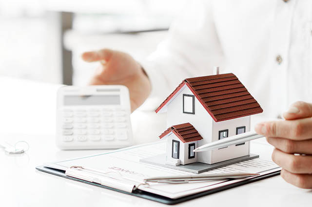 A person holds a pen, pointing at a small model house on a clipboard with documents, while using a calculator, suggesting real estate or mortgage planning.