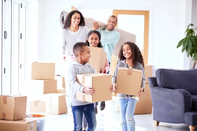 A smiling family of four carries boxes and a rolled-up rug into a bright, modern home, surrounded by moving boxes. The parents and two children appear happy and excited to be moving in. A smiling family of four carries boxes and a rolled-up rug into a bright, modern home, surrounded by moving boxes. The parents and two children appear happy and excited to be moving in.