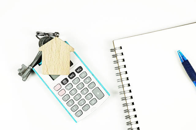 A calculator with keys and a house-shaped keychain, placed next to a spiral notebook and a blue pen on a white background. A calculator with keys and a house-shaped keychain, placed next to a spiral notebook and a blue pen on a white background.