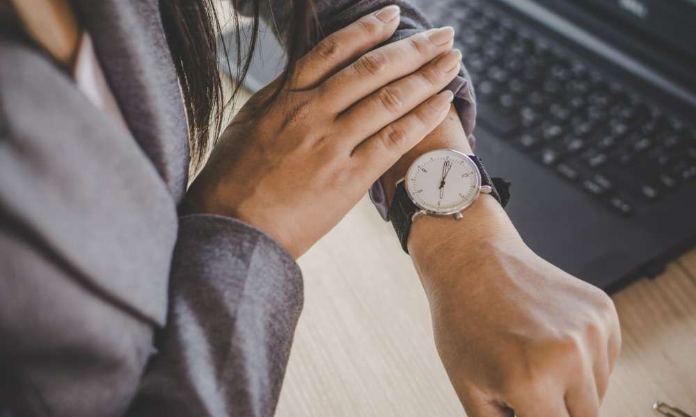 A person in a suit checks the time on their wristwatch while sitting at a desk next to a laptop. A person in a suit checks the time on their wristwatch while sitting at a desk next to a laptop.
