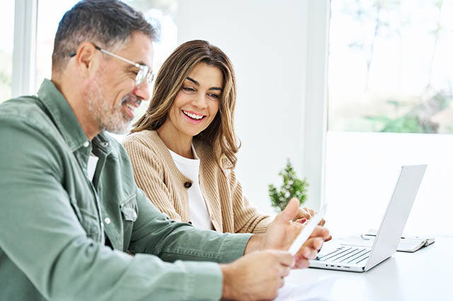 A man and woman sit at a desk with a laptop, smiling and looking at paperwork together in a bright, modern room.