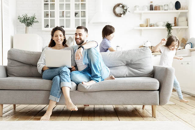 A smiling couple sits on a gray sofa using a laptop, while two children play in the bright, modern living room and kitchen in the background. The atmosphere is cozy and relaxed.