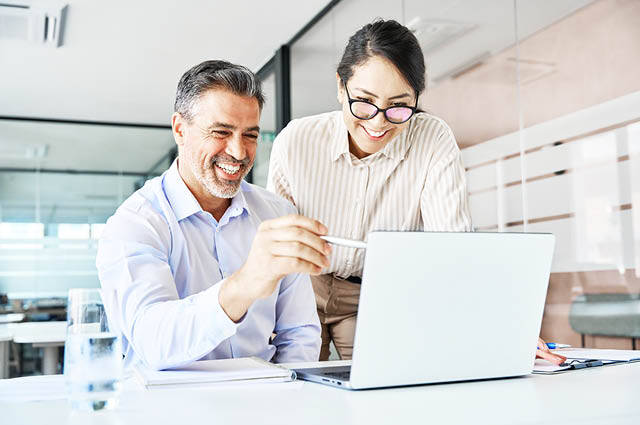 Two colleagues, a man and a woman, are smiling and looking at a laptop screen together in a bright, modern office. The man is holding a pen and pointing at the screen while the woman stands beside him wearing glasses. Two colleagues, a man and a woman, are smiling and looking at a laptop screen together in a bright, modern office. The man is holding a pen and pointing at the screen while the woman stands beside him wearing glasses.