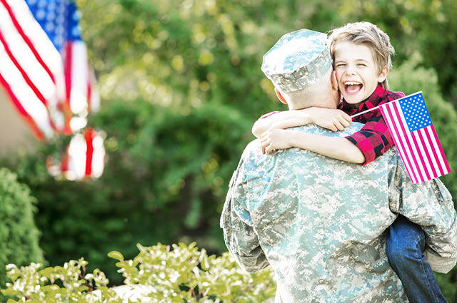 A soldier in uniform holds a smiling child who is holding a small American flag. An American flag is visible in the background, with greenery surrounding them.