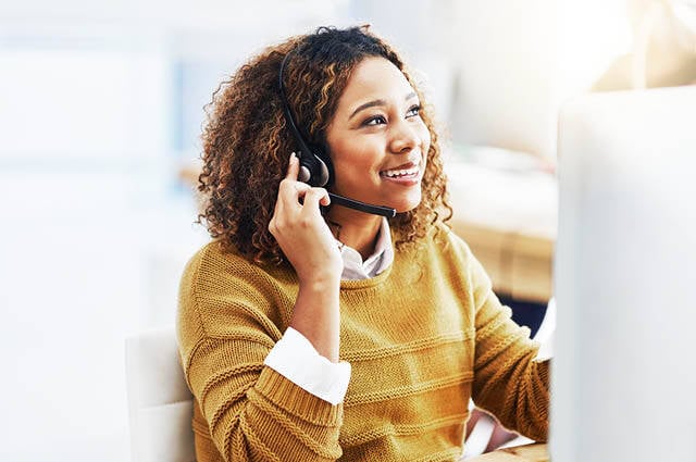 A woman with curly hair, wearing a mustard yellow sweater, sits at a desk and smiles while talking on a headset in a bright, modern office environment. A woman with curly hair, wearing a mustard yellow sweater, sits at a desk and smiles while talking on a headset in a bright, modern office environment.