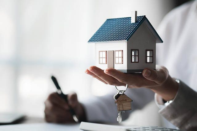 A person holds a small model house and keys in one hand while signing documents with the other hand, symbolizing real estate or home ownership. A person holds a small model house and keys in one hand while signing documents with the other hand, symbolizing real estate or home ownership.