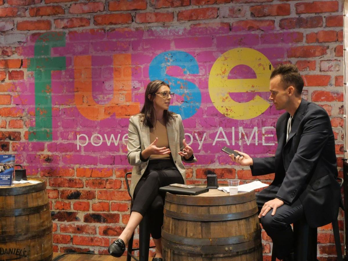 Two people sit on wooden barrels having a conversation in front of a colorful fuse powered by AIME sign on a brick wall. One person gestures while speaking, and the other holds a phone, listening attentively.