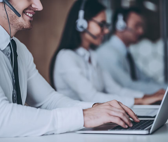 Three customer service representatives wearing headsets are seated in a row at desks, working on laptops and speaking with clients. The focus is on the foreground person’s hands typing on a laptop keyboard.