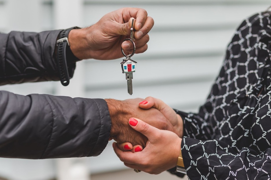 Two people shaking hands while one hands over a house key with a house-shaped keychain, symbolizing a real estate transaction or new home purchase.