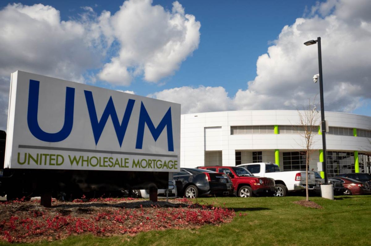 Large sign reading UWM United Wholesale Mortgage stands in front of a modern white building with green accents, a parking lot filled with vehicles, and a cloudy blue sky above. Large sign reading UWM United Wholesale Mortgage stands in front of a modern white building with green accents, a parking lot filled with vehicles, and a cloudy blue sky above.