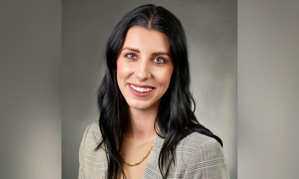 A woman with long, wavy black hair and green eyes smiles at the camera. She is wearing a light plaid blazer, a beige top, and a gold chain necklace, with a neutral gray background behind her.