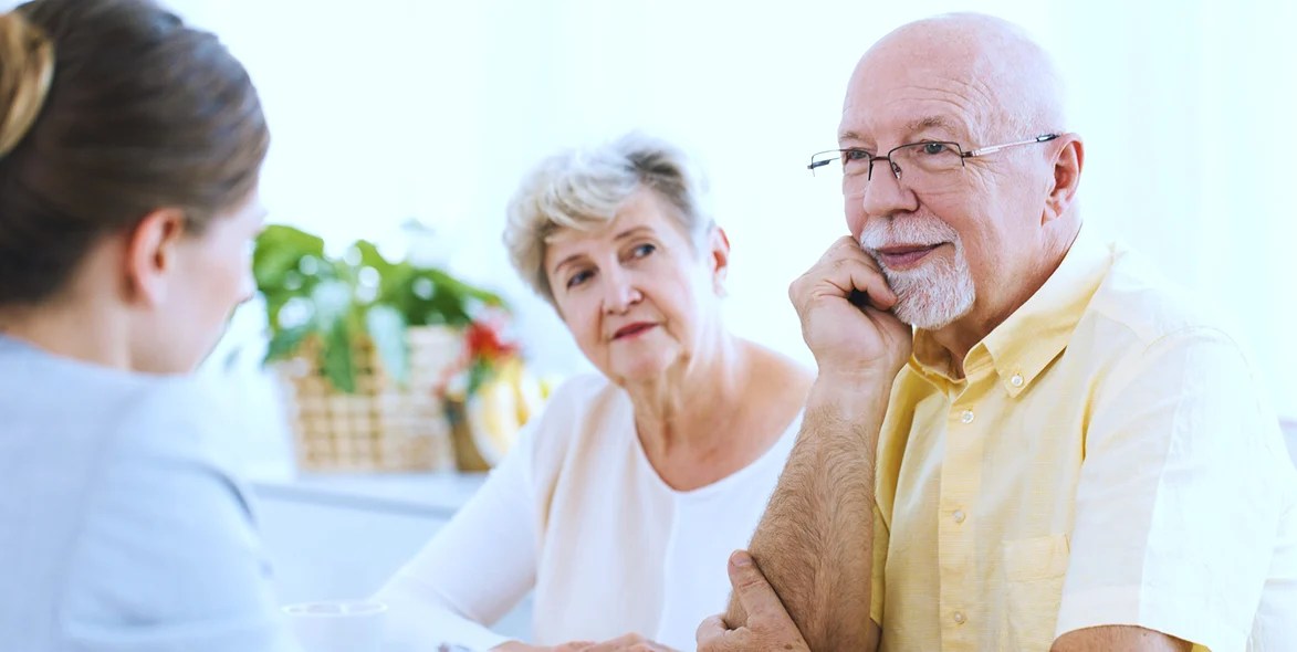 An elderly man and woman sit at a table, attentively listening to a younger woman. The older man wears a yellow shirt and glasses, while the woman wears white. They appear to be in a bright, homey setting. An elderly man and woman sit at a table, attentively listening to a younger woman. The older man wears a yellow shirt and glasses, while the woman wears white. They appear to be in a bright, homey setting.