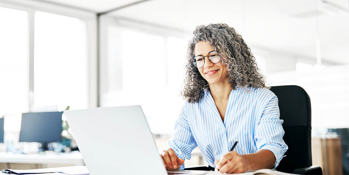 A woman with curly gray hair and glasses smiles while working on a laptop at a bright, modern office desk, holding a pen and surrounded by papers. A woman with curly gray hair and glasses smiles while working on a laptop at a bright, modern office desk, holding a pen and surrounded by papers.
