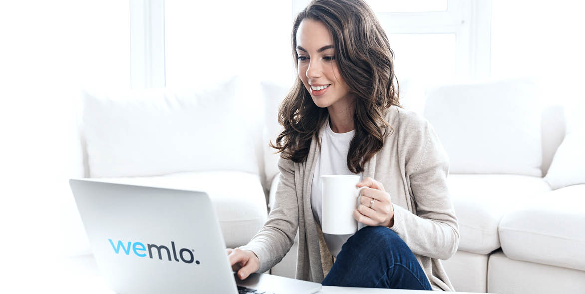 A woman sits on a white sofa, smiling and holding a mug in one hand while using a laptop with the wemlo logo on it. The background is bright and features large windows.