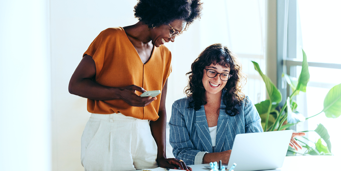 Two women smiling and working together in a bright office; one is sitting at a desk using a laptop, while the other stands beside her holding a smartphone. Both appear engaged and happy.