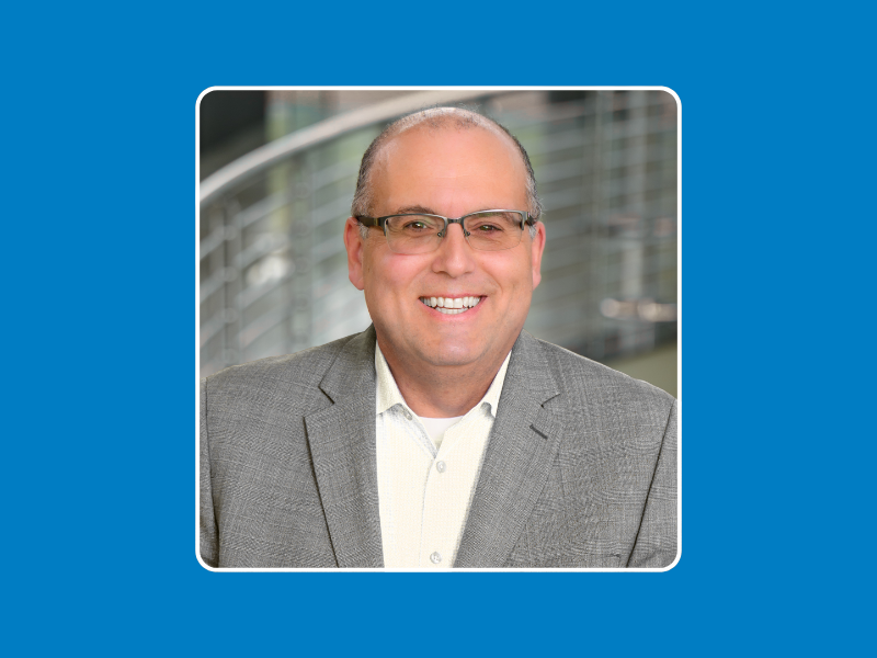 Man's headshot set against a blue background. Man is bald wearing glasses, a white collared shirt, and a grey blazer.