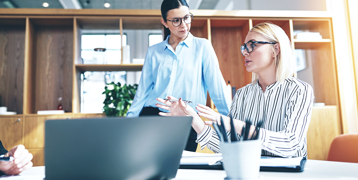 Two professionals in an office discussing paperwork and workflows, sitting at a desk with a laptop and documents, representing organization during busy mortgage seasons.