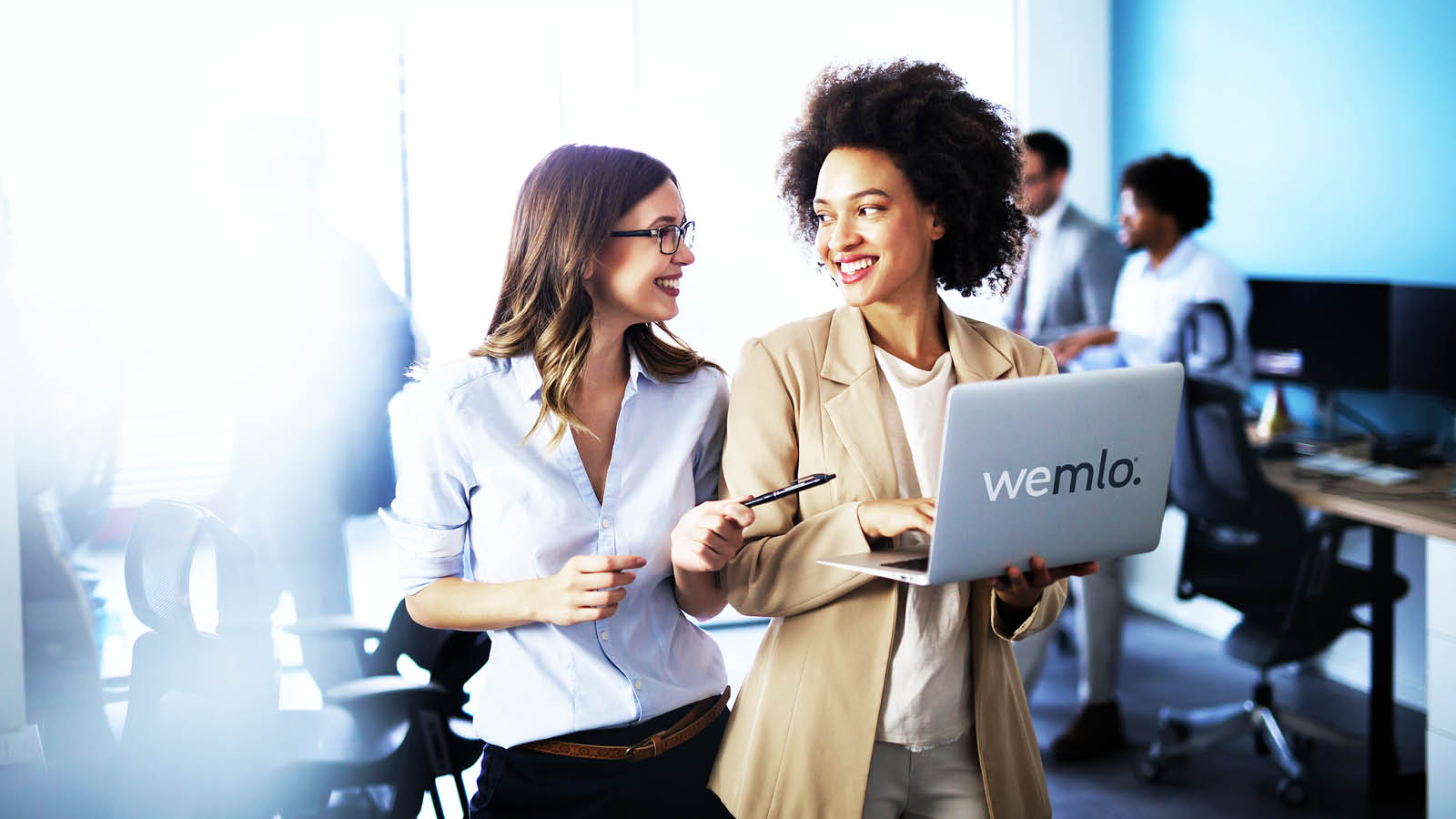Two professionals collaborating in a modern office while reviewing information on a laptop, with additional team members working in the background.
